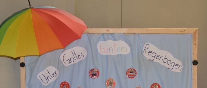 Eine Ausstellung mit einem farbenfrohen Regenschirm, blauem Tuch und einem Holzrahmen. Es zeigt viele bunte Kreise und Texte wie buntem und Regenbogen. Unten steht ein Tisch mit verschiedenen Gegenständen, einschließlich Flaschen und anderen Gegenständen in Plastik.