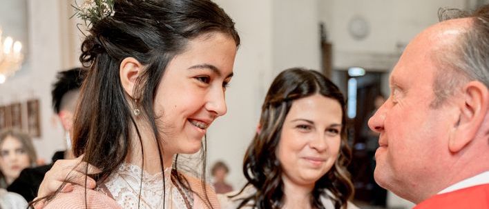 Ein Mann in einem roten Gewand spricht mit einer Frau in traditioneller Kleidung, während eine andere Frau im Hintergrund zuschaut. Die Szene spielt sich in einem kirchenähnlichen Ambiente ab.