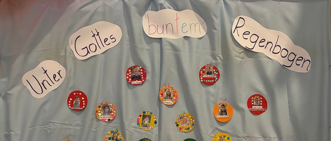 Ein Klassenzimmer-Bulletinbrett mit grauem Hintergrund zeigt kreisförmige Bilder von Schülern mit ihren Namen und kleinen Wolken mit Wörtern darüber. Die Wolken lesen 'Gottes' und 'Regenbogen'.