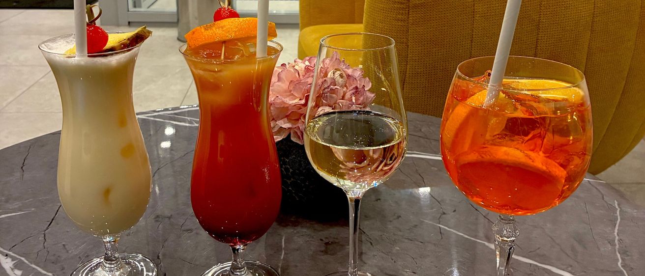 Vier Gläser mit verschiedenen Getränken stehen auf einem Marmortisch. Eines hat eine Kirsche und eine Banane obenauf. Ein anderes hat eine Orangenscheibe. Das dritte hat eine Blume. Das vierte Glas hat Orangensaft. Ein gelber Sessel steht dahinter.