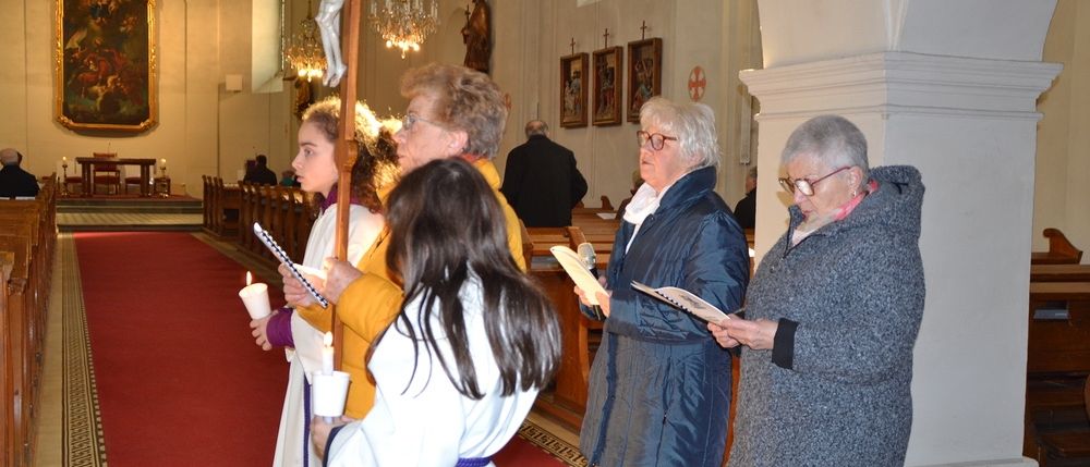 In einer Kirche stehen mehrere Personen mit Kerzen. Sie tragen verschiedene Kleidungsstücke, einschließlich weißer Gewänder und Brillen. Ein Kruzifix ist an einem Pfahl montiert.