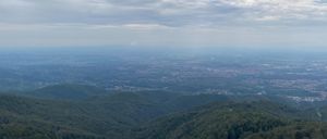 Ein Panoramablick auf eine bergige Landschaft mit dichten grünen Wäldern und einer entfernten Stadtlandschaft unter einem bewölkten Himmel.