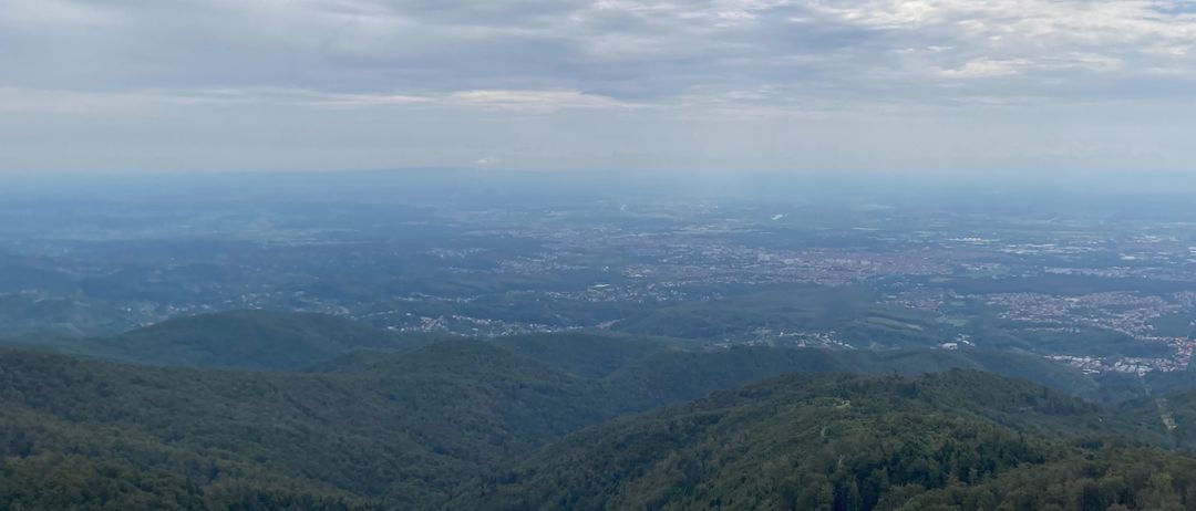 Ein Panoramablick auf eine bergige Landschaft mit dichten grünen Wäldern und einer entfernten Stadtlandschaft unter einem bewölkten Himmel.