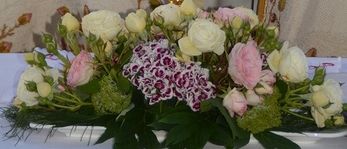 Ein Priester steht an einem mit Blumen geschmückten Altar, hält ein Mikrofon in der Hand, mit einem jungen Mädchen im Hintergrund.