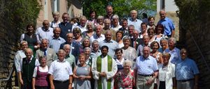 Eine große Gruppe älterer Menschen, einschließlich eines Priesters, steht auf den Stufen vor einem Gebäude, umgeben von viel Grün und einem klaren Himmel.
