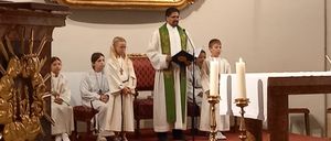 Ein Priester steht mit Kerzen am Altar und liest Kindern vor, die in weißen Gewändern sitzen. Ein großes Gemälde hängt hinter ihm an einer Wand.