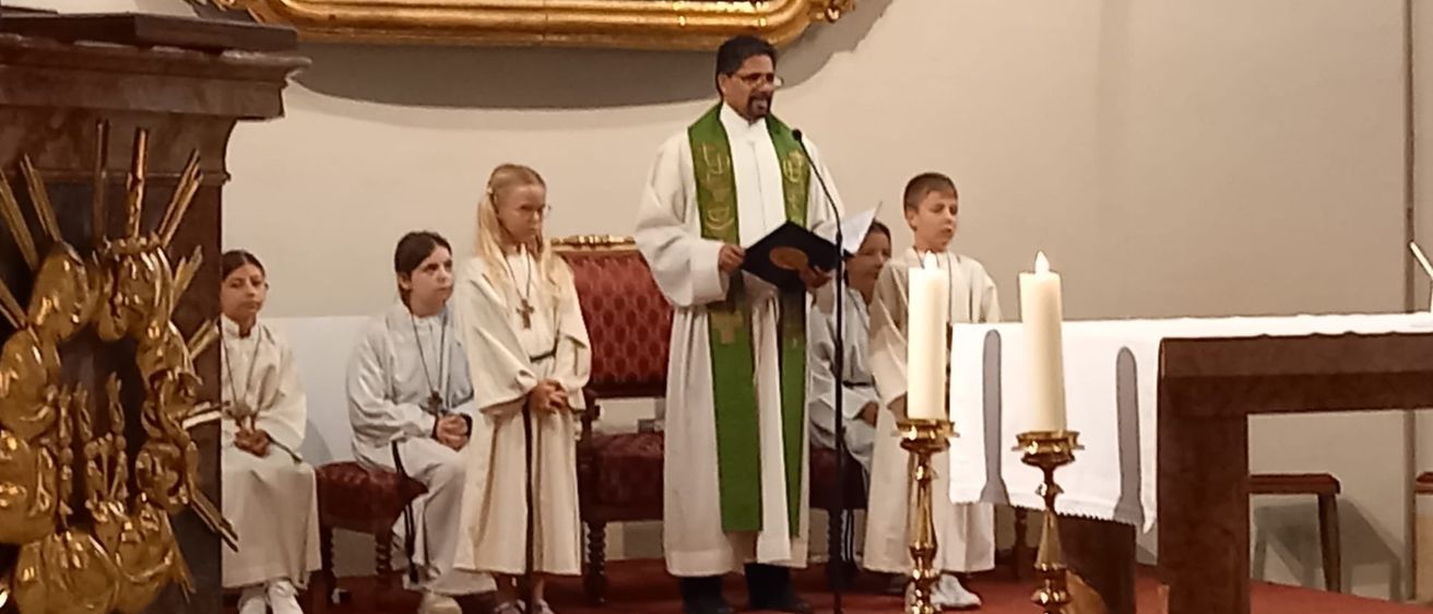 Ein Priester steht mit Kerzen am Altar und liest Kindern vor, die in weißen Gewändern sitzen. Ein großes Gemälde hängt hinter ihm an einer Wand.