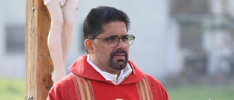 Ein Priester mit Brille und roter Robe hält ein Buch und ein Mikrofon in der Hand und steht auf einem Feld.