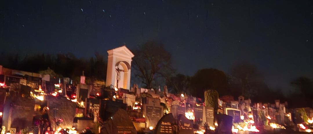 Ein Friedhof, der bei Nacht von Kerzen und Lichtern erleuchtet wird, mit einer Kapelle und Bäumen im Hintergrund.