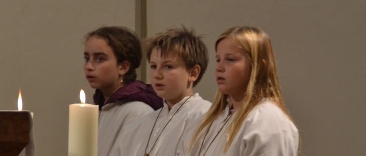 Drei Kinder in weißen Gewändern stehen neben einer Kerze in einer Kirche. Ein Kind trägt eine karmesinrote Kapuze.