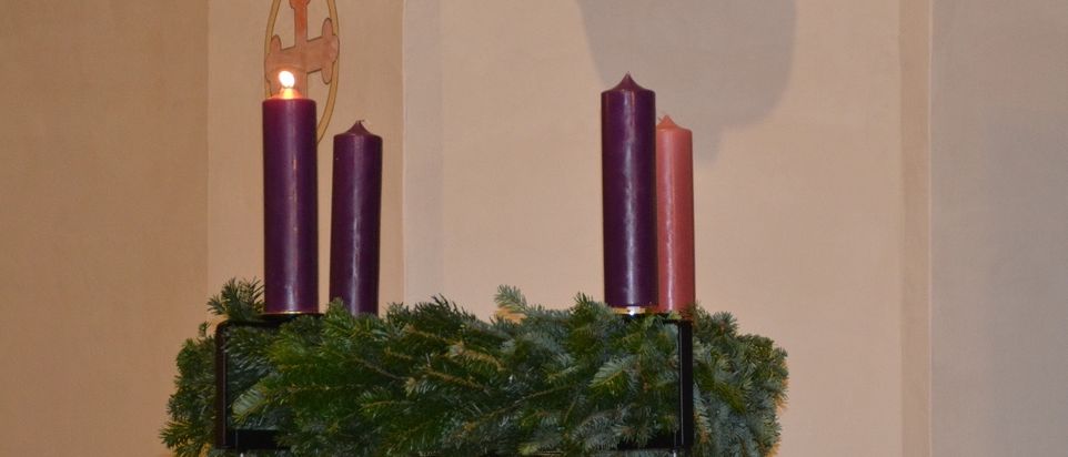 Ein Kerzenständer mit vier brennenden Kerzen, drei lila und eine rosa, ist mit grünem Laub geschmückt. Dahinter befindet sich eine beige Wand mit einem goldenen Kreuzsymbol.