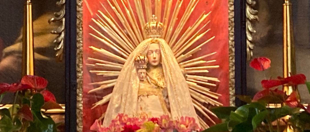 Ein religiöser Altar mit einem verzierten Rahmen zeigt ein Bild der Jungfrau Maria, umgeben von roten Blumen. Sechs Kerzen sind angezündet und um den Rahmen platziert.