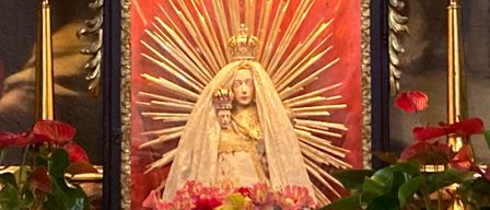 Ein religiöser Altar mit einem verzierten Rahmen zeigt ein Bild der Jungfrau Maria, umgeben von roten Blumen. Sechs Kerzen sind angezündet und um den Rahmen platziert.