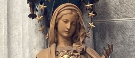 Eine Statue einer weiblichen Figur mit einem Schleier und einer Lilienzweig in der Hand, mit einem goldenen Herzen auf der Brust.