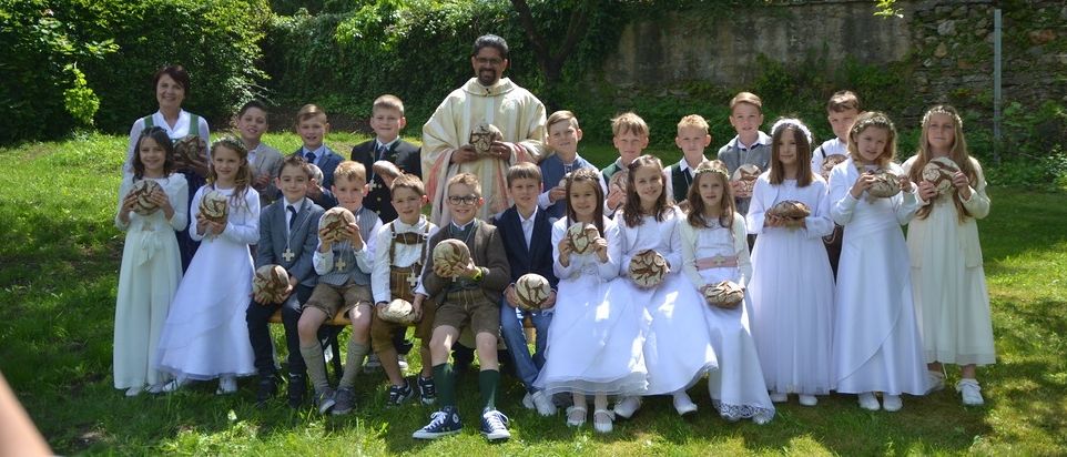 Eine Gruppe von Kindern in weißen Kleidern und Jungen in Anzügen versammelt sich mit einem Priester in einem Garten und hält dekoratives Brot in den Händen.