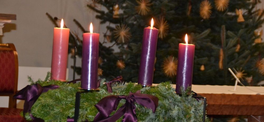 Vier lila Kerzen brennen auf einem Kranz vor einem geschmückten Weihnachtsbaum.