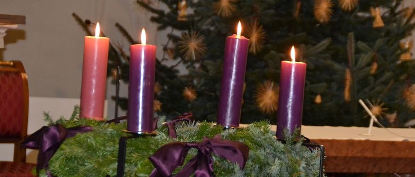 Vier lila Kerzen brennen auf einem Kranz vor einem geschmückten Weihnachtsbaum.