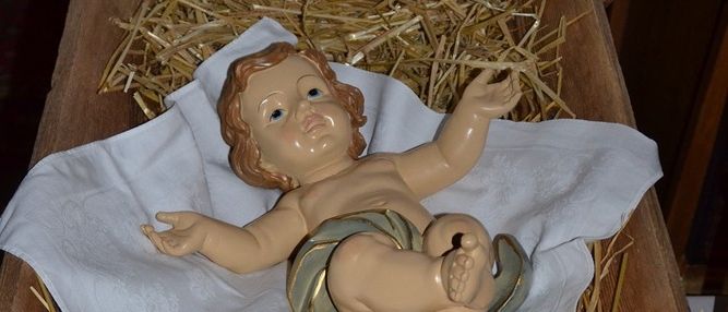 Eine kleine, detaillierte Figur des Baby Jesus ruht in einem hölzernen Krippenbett mit Stroh, unter einem weißen Spitzentuch. Ein weißes Tuch bedeckt teilweise das Krippenbett.