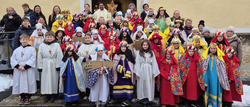 Eine Gruppe von Kindern und Erwachsenen in farbenfrohen Kostümen und Hüten posiert für ein Foto vor einem Gebäude. Einige halten Stäbe und Kronen.
