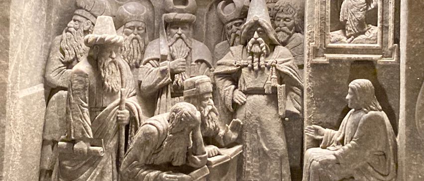 Eine Steinschnitzerei zeigt eine Gruppe von Männern, einige mit Hüten, die um eine sitzende Figur herum sitzen und stehen. Die sitzende Figur ist von anderen umgeben, einige halten Gegenstände.