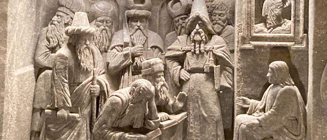 Eine Steinschnitzerei zeigt eine Gruppe von Männern, einige mit Hüten, die um eine sitzende Figur herum sitzen und stehen. Die sitzende Figur ist von anderen umgeben, einige halten Gegenstände.