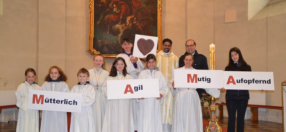 Eine Gruppe von Kindern in weißen Gewändern steht auf Stufen und hält Schilder mit deutschen Wörtern. Priester und andere Erwachsene stehen dahinter. Kerze auf dem Altar, Gemälde an der Wand.