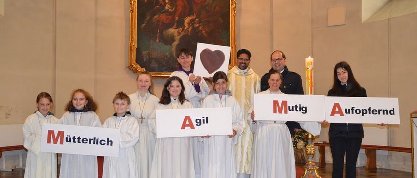 Eine Gruppe von Kindern in weißen Gewändern steht auf Stufen und hält Schilder mit deutschen Wörtern. Priester und andere Erwachsene stehen dahinter. Kerze auf dem Altar, Gemälde an der Wand.
