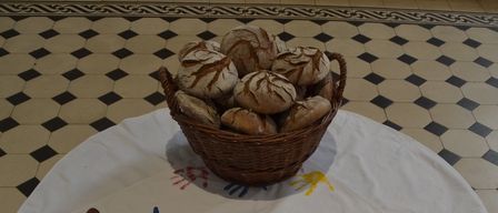 Ein runder Tisch, bedeckt mit einem weißen Tuch, das farbenfrohe Fischzeichnungen und einen Korb mit Brot aufweist. Der Boden ist mit schwarzen und weißen Quadraten gefliest.