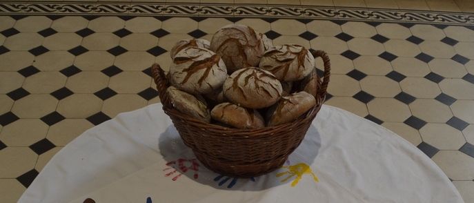 Ein runder Tisch, bedeckt mit einem weißen Tuch, das farbenfrohe Fischzeichnungen und einen Korb mit Brot aufweist. Der Boden ist mit schwarzen und weißen Quadraten gefliest.