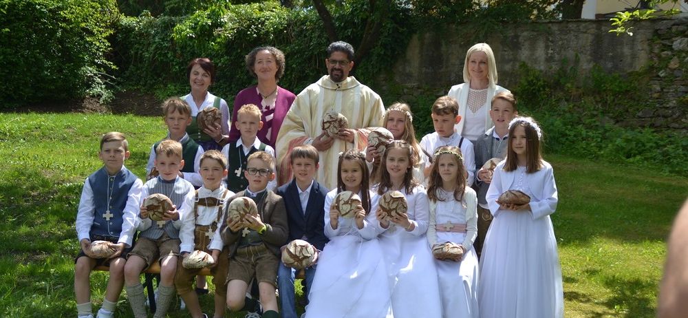 Eine Gruppe von Kindern und Erwachsenen posiert für ein Foto in einem Garten mit Brot in den Händen. Ein Priester steht unter ihnen.