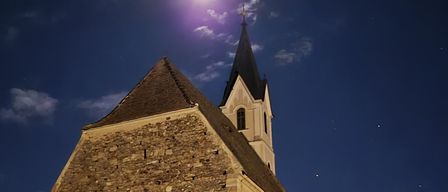 A night view of an old stone church with a steeple and a moon illuminating it. The sky is blue with scattered clouds.