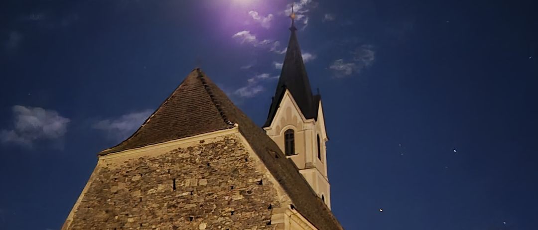 Ein Nachtblick auf eine alte Steinkirche mit einem Turm und einem Mond, der sie erhellt. Der Himmel ist blau mit verstreuten Wolken.