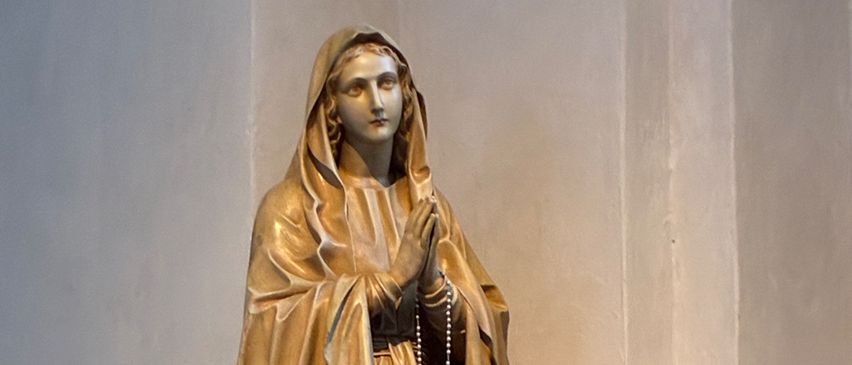 A golden statue of a woman, possibly the Virgin Mary, is praying. She is holding a rosary in her hands and is wearing a brown robe. There are flowers on the ground in front of her.