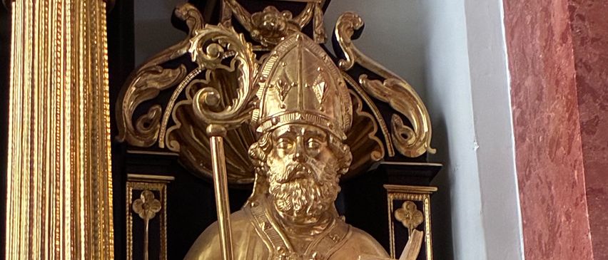 A golden statue of a bishop holding a book and a staff, adorned with intricate designs, stands on a marble pedestal.