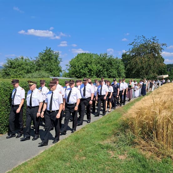 Eine Reihe uniformierter Männer läuft auf einem Weg mit einem grünen Hecken- und Grasfeld auf einer Seite und einem Weizenfeld auf der anderen. Bäume und Büsche sind im Hintergrund unter einem klaren Himmel.