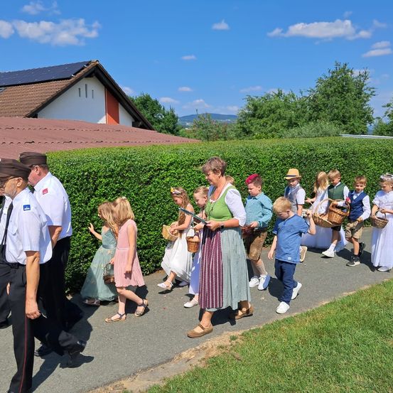 Eine Gruppe von Kindern und Erwachsenen geht einen Gehweg entlang und trägt Körbe in einer sonnigen Umgebung.