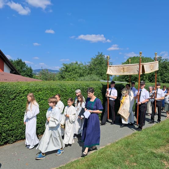 Eine Gruppe junger Menschen in weißen Gewändern geht hinter einem von Erwachsenen gehaltenen Baldachin an einem sonnigen Tag.