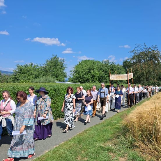 Eine Gruppe von Menschen in traditioneller Kleidung, einige mit Papieren in der Hand, gehen in einer Reihe auf einem Pfad mit einem Banner im Hintergrund.