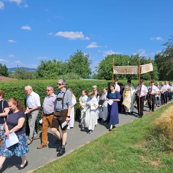 Eine Gruppe von Menschen in traditioneller bayerischer Kleidung geht auf einem Weg mit einem Baldachin, umgeben von Hecken und Grün.