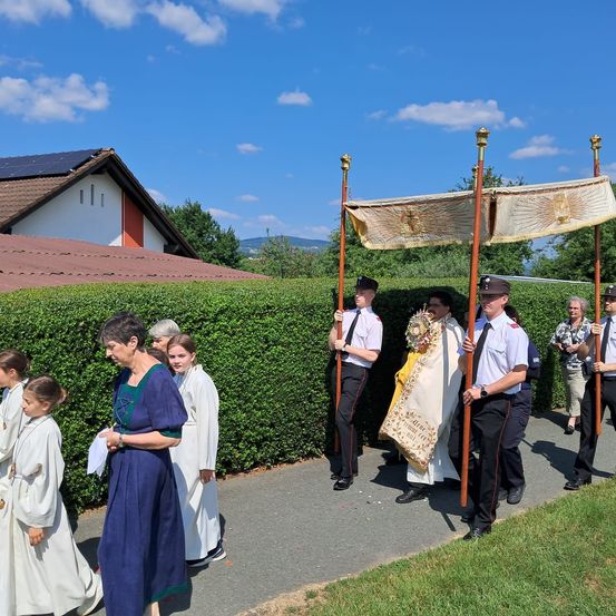 Eine Prozessionsgruppe geht auf einem Pfad, begleitet von uniformierten Personen. Ein Priester steht in der Mitte, flankiert von anderen in weißen Gewändern. Hinter ihnen wird ein Baldachin von zwei Personen gehalten. Die Szenerie ist vor einem Haus mit Solarpanelen und einem Heckenhintergrund.