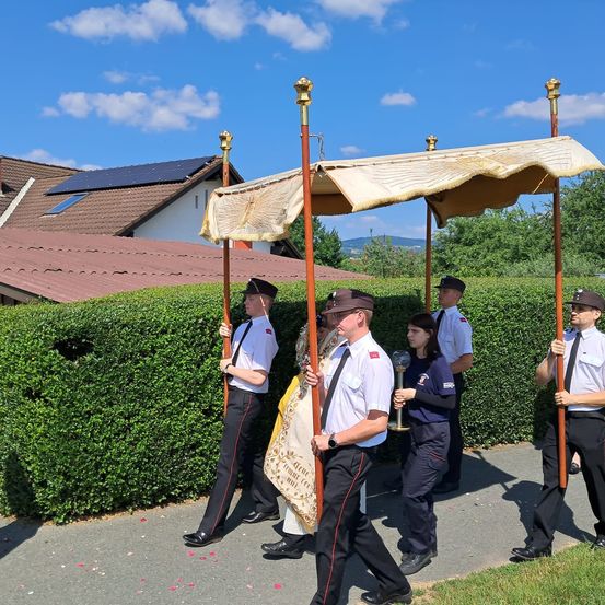 Eine Gruppe uniformierter Personen, darunter eine Frau mit einem Kelch, geht unter einem Baldachin, möglicherweise während einer Prozession. Sie befinden sich auf einem Weg, der von Hecken gesäumt ist. Hinter ihnen steht ein Haus mit Solarmodulen auf dem Dach.