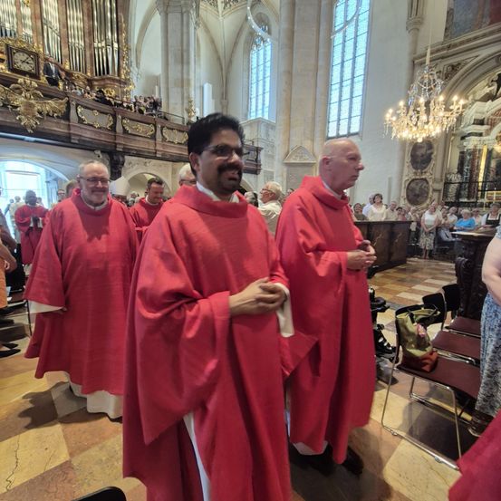 Drei Priester in roten Gewändern stehen in einer Kirche, möglicherweise während einer Zeremonie. Sie stehen mit gefalteten Händen der Zuschauer zugewandt.