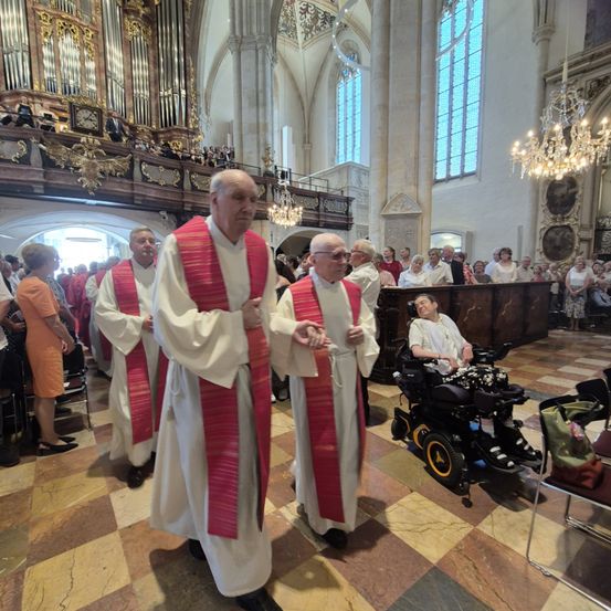 Eine Gruppe von Priestern in traditionellen Gewändern geht durch eine Kathedrale. Eine Frau im Rollstuhl beobachtet vom Gang aus.
