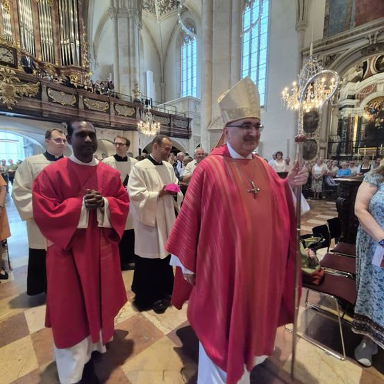Eine Gruppe von Priestern in roten und weißen Gewändern steht in einer Kirche. Einer hält einen Stab und trägt die Bischofsmütze. Eine Menge beobachtet im Hintergrund.
