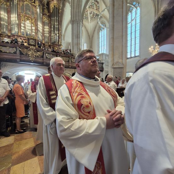 Priester stehen in einer Kirche, einer mit Brille und einer roten Stola, ein anderer in einem weißen Gewand, stehen sich gegenüber mit gefalteten Händen. Hinter ihnen stehen und sitzen Menschen, mit einer verzierten Orgel darüber.