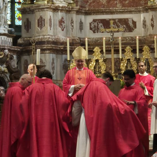 Eine Gruppe von Priestern in roten Roben versammelt sich um einen Bischof, der eine Mitra und eine Brille trägt. Sie befinden sich in einer Kirche mit Kerzen und einem Kruzifix.