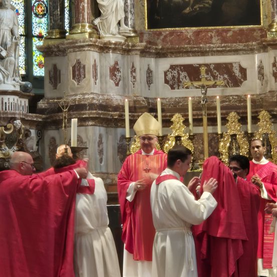 Ein Priester steht vor einem Altar, umgeben von anderen Geistlichen in roten und weißen Gewändern, in einer Kathedrale. Kerzen sind angezündet, und Statuen schmücken die Wände.