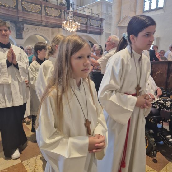 Eine Gruppe von Kindern in weißen Gewändern steht in einer Kirche, möglicherweise an einer religiösen Zeremonie teilnehmend. Die Kinder schauen nach vorne, und eines von ihnen hält ein Kreuz. Dahinter stehen und sitzen verschwommene Menschen, möglicherweise im Gebet. Eine Kronleuchter hängt von der Decke.