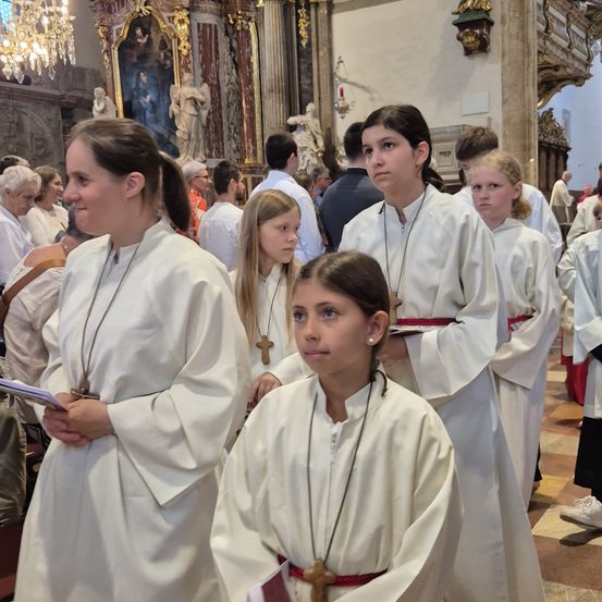Eine Gruppe junger Mädchen in weißen Gewändern, wahrscheinlich während einer religiösen Zeremonie, steht in einer Kirche. Sie tragen Halsketten mit Kreuzen und roten Schärpen. Dahinter sind ältere Personen anwesend.