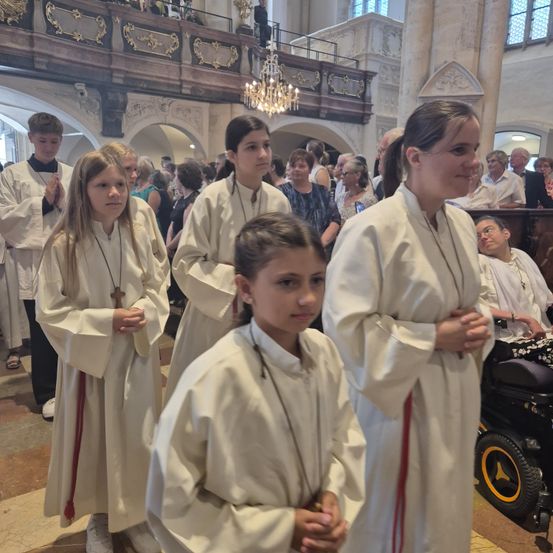 Eine Gruppe von Kindern in weißen Gewändern betet in einer Kirche. Sie stehen neben einer lächelnden erwachsenen Frau. Dahinter sitzen mehrere Menschen auf den Kirchenbänken.
